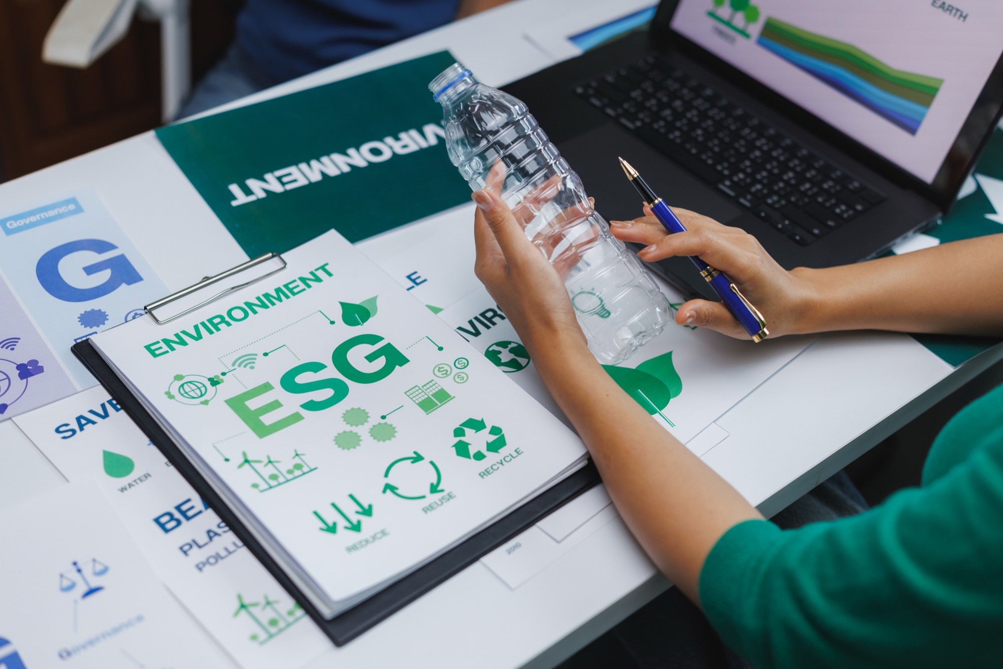 Hands reviewing an ESG report bound with environmental icons next to a laptop